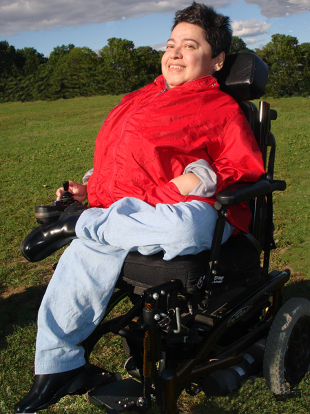 woman with short hair in powerchair, with red jacket, crossed legs and foxy smile